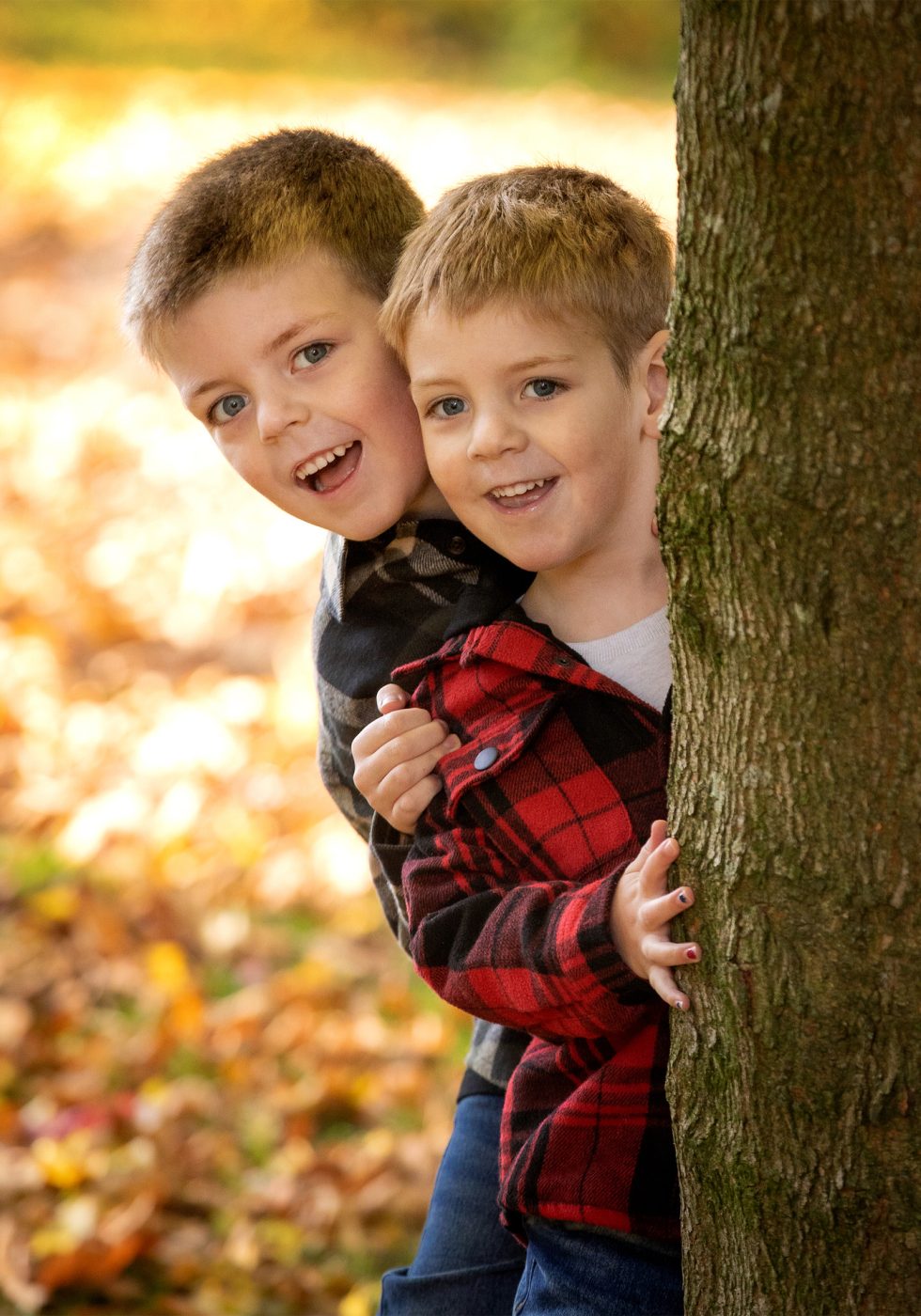 Two young boys with short light brown hair smile and peek from behind a tree in an autumn park. They wear checked jackets, one red and one grey. The ground is covered with colourful fallen leaves, and the background glows with soft, warm sunlight.