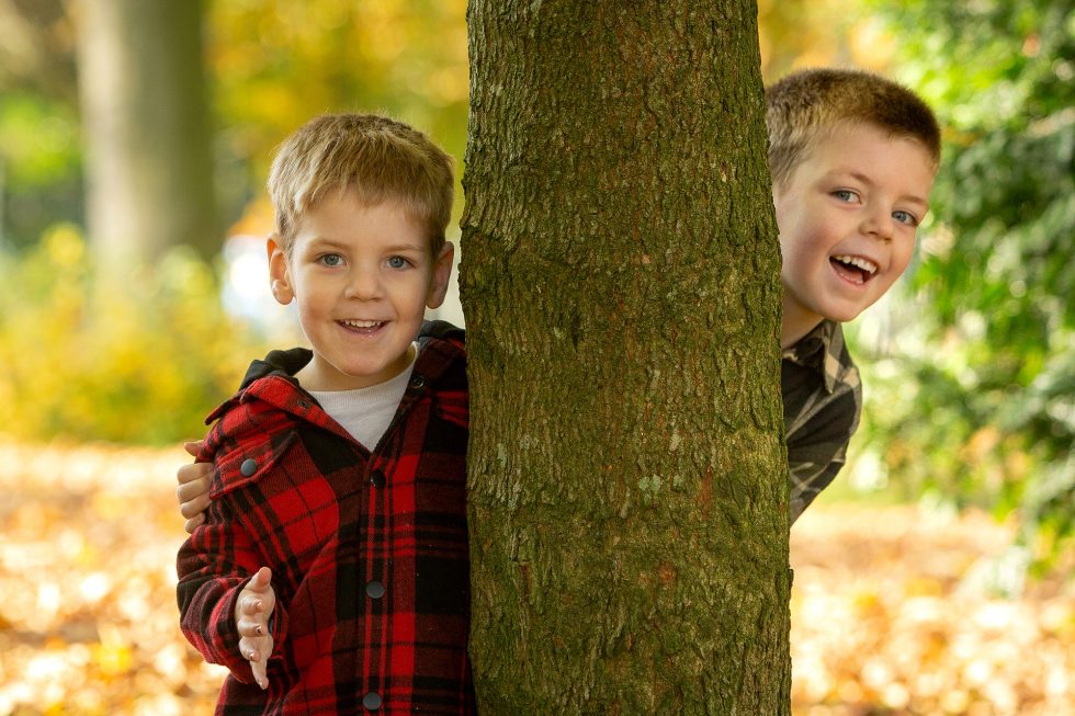 Two young boys smile and play behind a tree in an autumn park. One boy in a red checked jacket stands in front, while the other in a brown checked shirt peeps from behind the tree, both surrounded by fallen yellow leaves and blurred foliage.