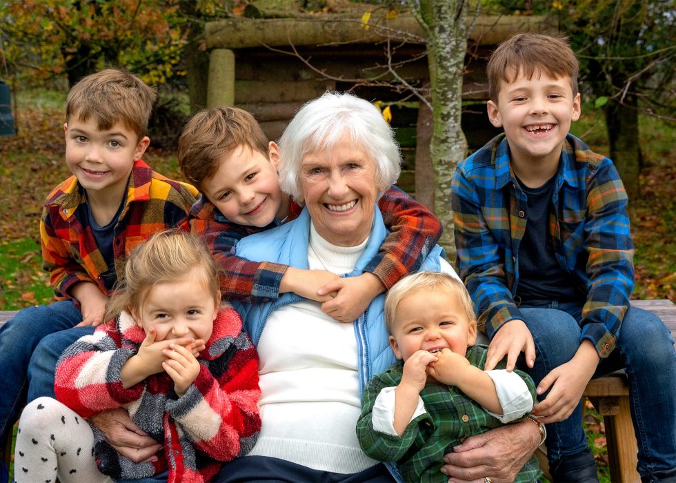 An elderly woman with short white hair sits outdoors, smiling and hugging five young children around her. The kids, three boys and two girls, are dressed in colourful checked shirts and jackets, smiling happily amid an autumnal background with trees and leaves.