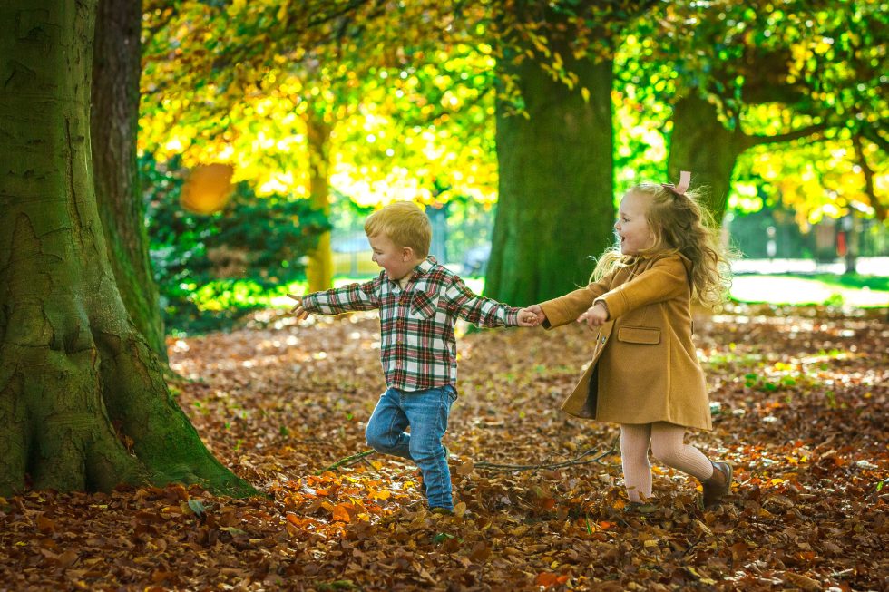 Two young children, a boy in a checked shirt and jeans and a girl in a brown coat and dress, run hand in hand through fallen autumn leaves in a sunlit park. Tall trees with green and yellow leaves frame the playful scene.