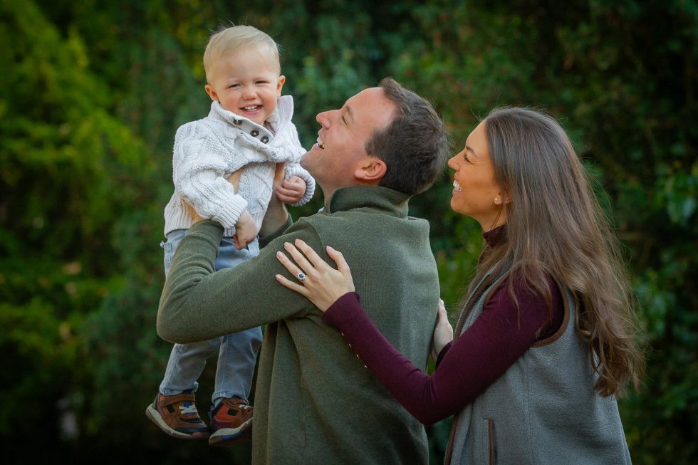 A man lifts a smiling toddler boy in a white jumper as a woman with long brown hair stands beside them, smiling. They are outdoors with green trees in the background, all wearing warm, casual clothes.