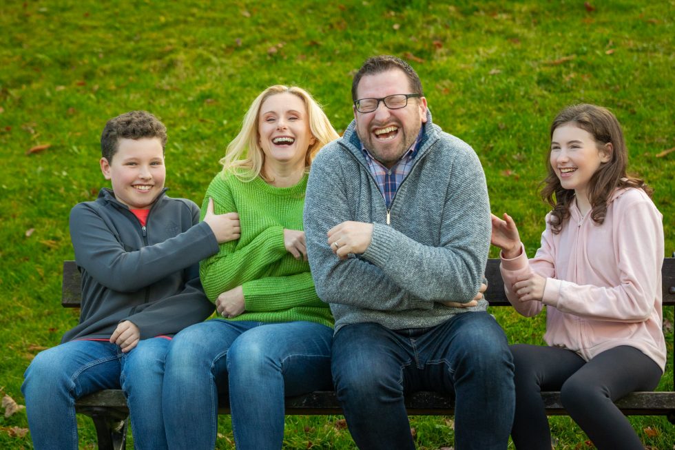 Four people sit close together on a bench outdoors, laughing and smiling. The group includes a woman in a green jumper, a man in a grey jumper, a boy in a grey zip-up, and a girl in a pink jacket. Green grass covers the background.
