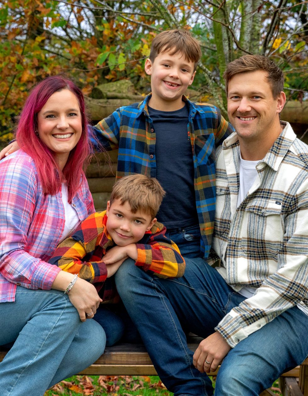 A family of four poses outdoors on a bench. The mother, with red hair, and the father, with light brown hair, both wear checked shirts and jeans. Their two young sons, also in checks, smile; one stands behind and the other sits, leaning on his mother’s knee. Autumn trees are behind them.