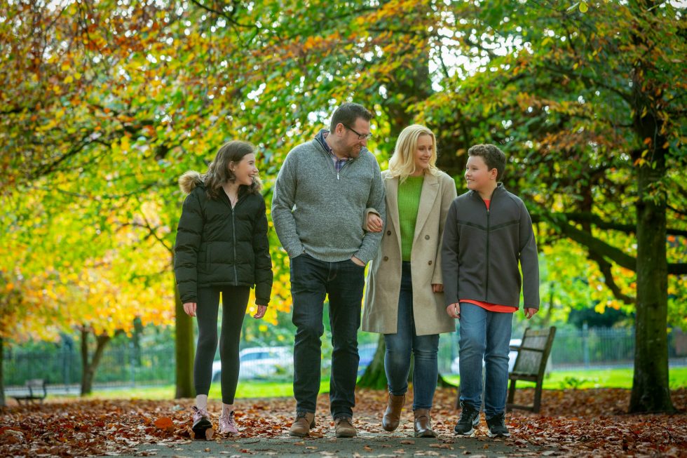 A family of four walks together in a park with autumn leaves on the ground. Two adults (a man and a woman) are in the centre, with a girl on the left and a boy on the right. Trees with colourful autumn foliage and a wooden bench are in the background.