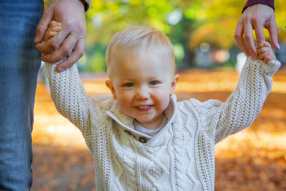 A smiling toddler with blond hair, wearing a cream knitted jumper, holds hands with two adults. The scene is outdoors in a sunlit park with autumn leaves on the ground and blurred trees in the background.