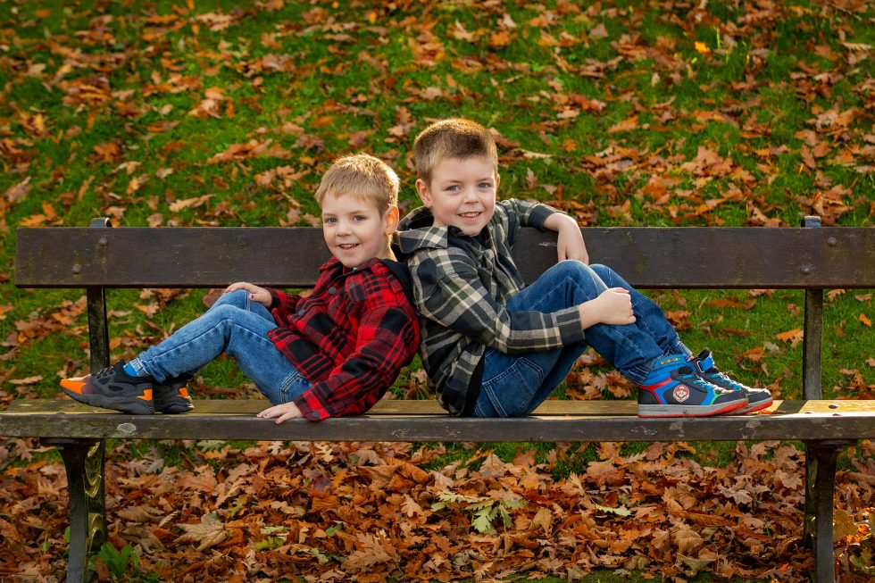 Two young boys sit back-to-back on a wooden bench outdoors, smiling. Both wear checked shirts and jeans. Fallen autumn leaves cover the ground, with green grass in the background. The scene is bright and cheerful.