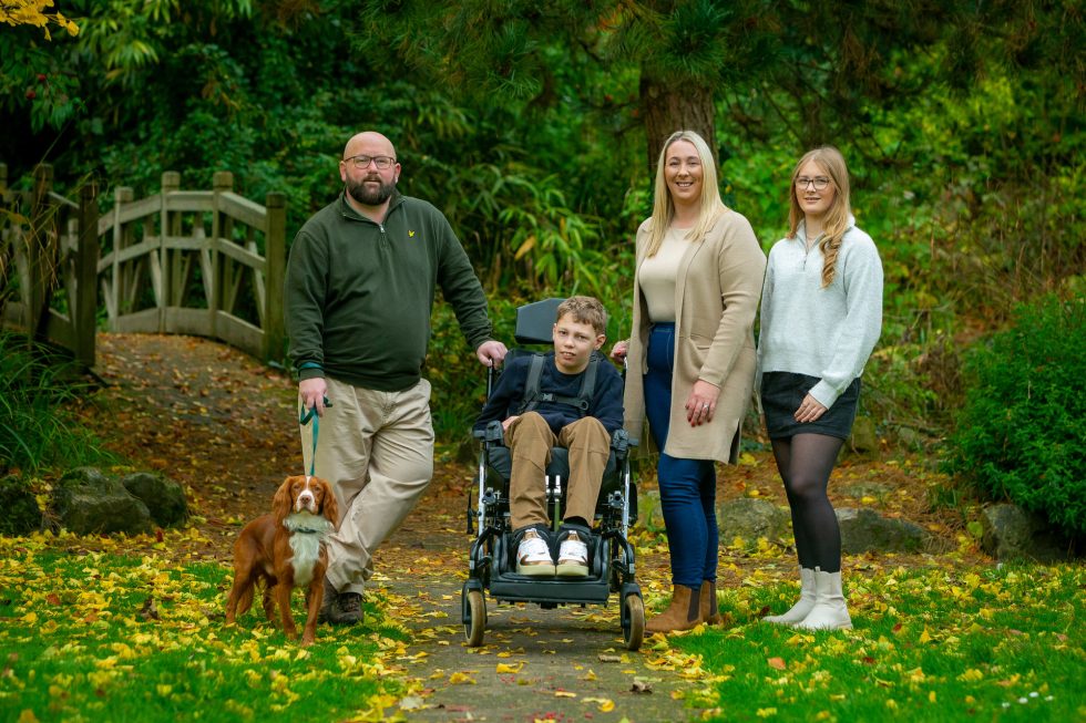 A family of four and a dog stand on a leaf-covered path in a green park. The boy in the centre sits in a wheelchair, flanked by two women and a man holding the dog’s lead. A wooden footbridge and lush foliage fill the background.