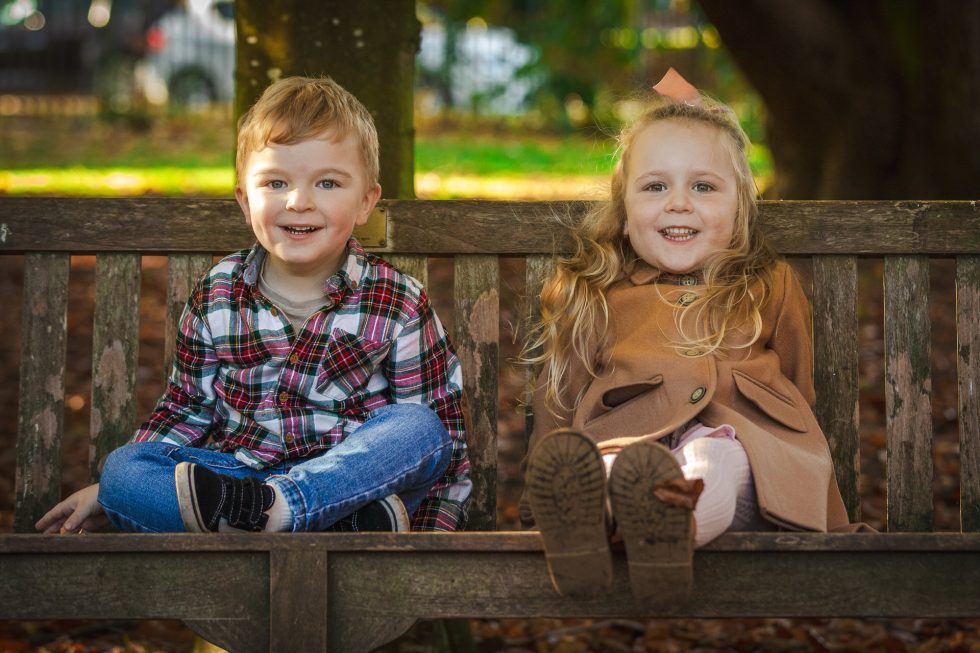 Two young children sit on a wooden bench outdoors. The boy on the left wears a checked shirt and jeans, smiling with legs crossed. The girl on the right wears a tan coat and pink bow, also smiling, with her legs stretched out and shoes visible. Autumn leaves are scattered below.