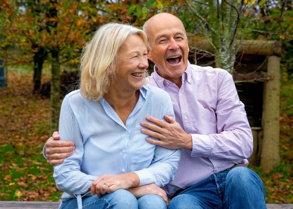 An older couple sit close together on a bench outdoors, laughing and smiling. The woman has blonde hair and wears a light blue shirt, while the man is bald and wears a pale striped shirt. Autumn leaves and trees fill the background, creating a warm, cheerful scene.