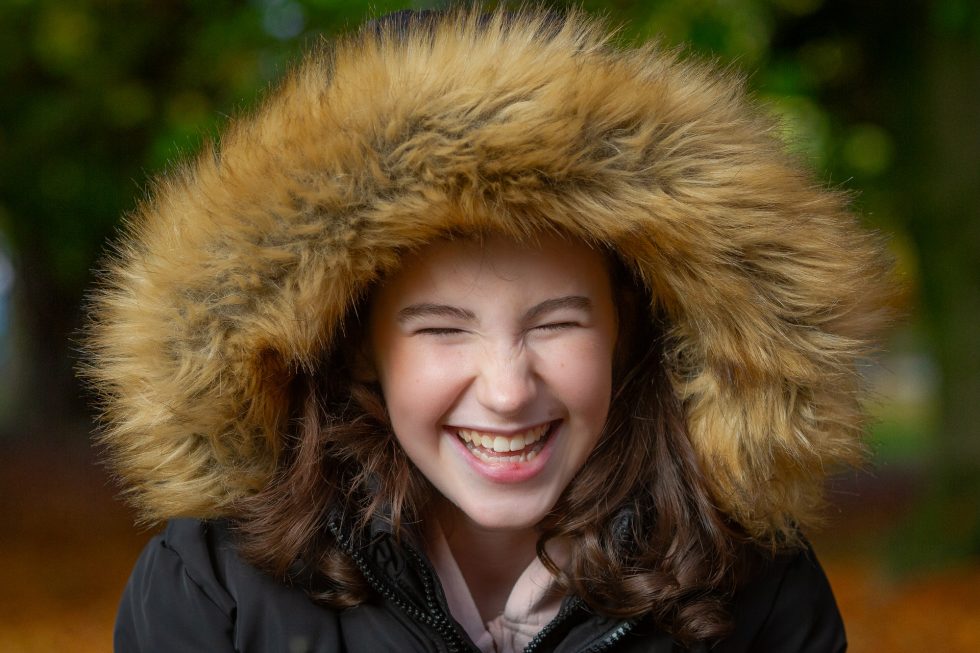 A young girl with long brown hair smiles broadly, eyes closed, wearing a dark coat with a large, fluffy, tan fur-lined hood. The background is softly blurred with hints of greenery and autumn colours. The overall mood is joyful and warm.