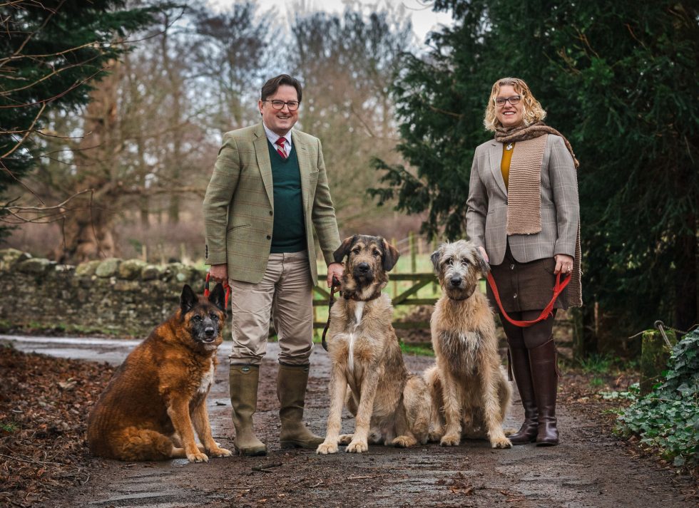 A man and woman stand on a path outdoors, smiling and holding leads of three large dogs. The man wears a green jumper, tweed jacket, tie, and boots; the woman wears a tan jacket, scarf, and brown skirt. Trees and a stone wall are in the background.