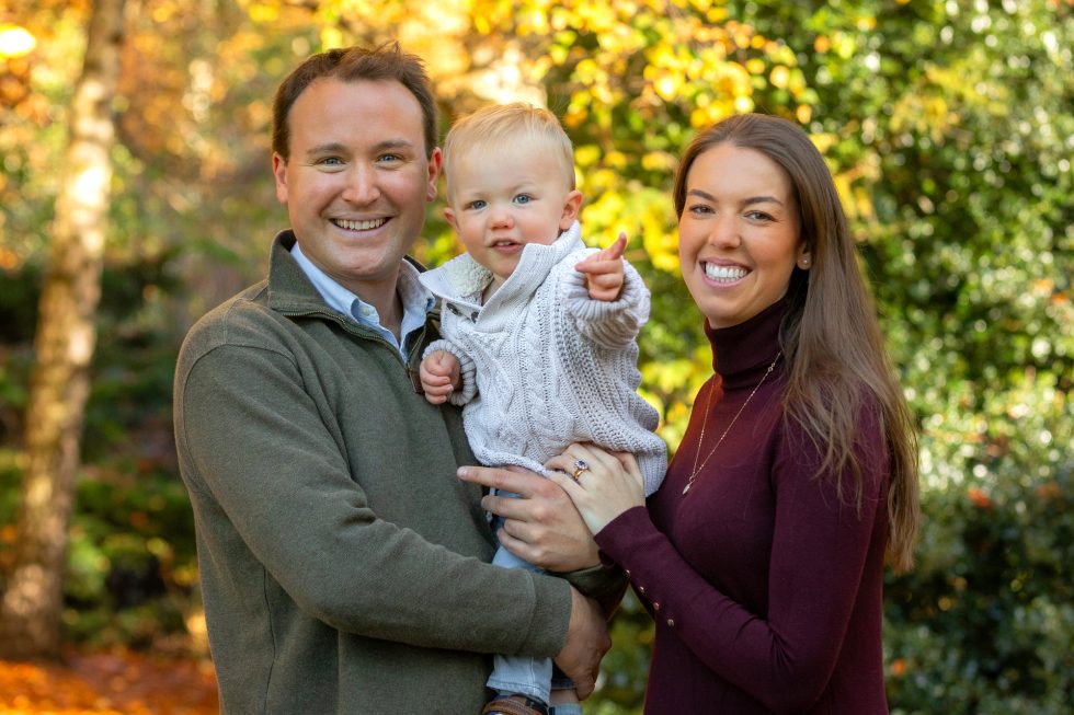 A smiling man and woman stand outdoors holding a baby boy. The baby is wearing a light jumper and points forward. The adults are dressed in autumn jumpers. Autumn foliage with green and yellow leaves fills the background. All appear happy and cheerful.