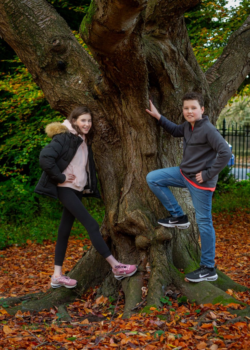 Two kids pose at the base of a large tree with rough bark. The girl on the left wears a black jacket, pink hoodie, black leggings, and pink trainers. The boy on the right wears a grey jacket, blue jeans, and black trainers. Fallen autumn leaves cover the ground.