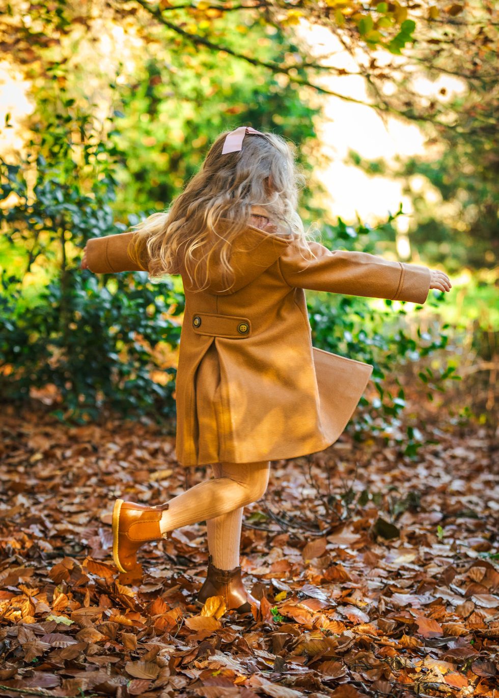 A young girl with long blonde hair, wearing a tan coat, tan tights, brown boots, and a light pink hair bow, spins joyfully among fallen autumn leaves in a sunlit, leafy outdoor setting, with her arms outstretched.