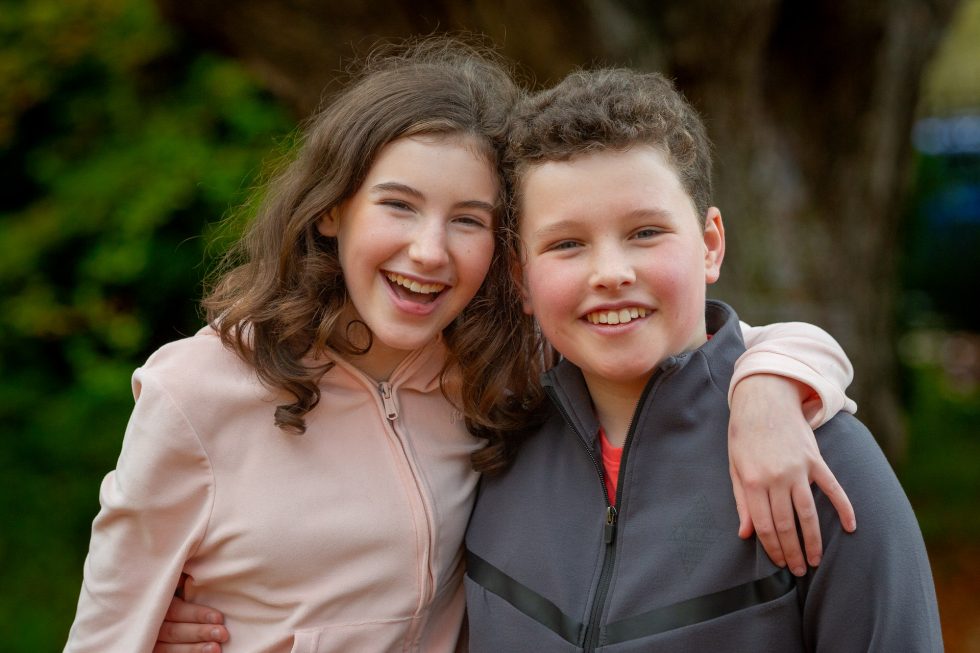 Two smiling children stand outdoors, one girl and one boy, both with curly brown hair. The girl, in a pale pink jacket, has her arm round the boy, who wears a grey jacket. They look at the camera with happy expressions, trees blurred in the background.