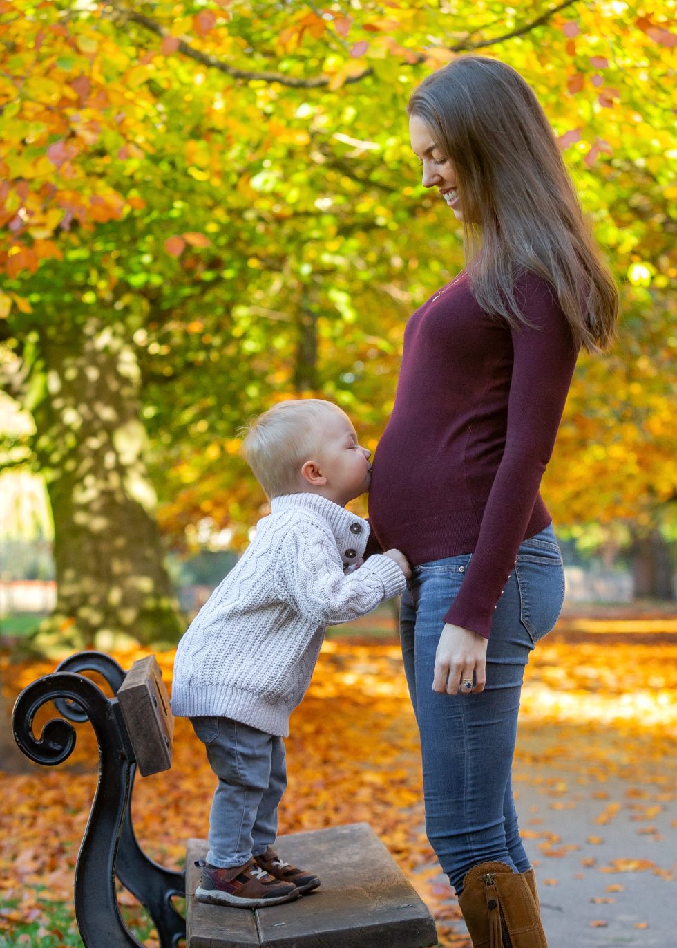 A young boy in a white jumper stands on a park bench, kissing the belly of a smiling pregnant woman with long brown hair, wearing a maroon jumper and jeans. They are outdoors surrounded by colourful autumn leaves and trees.