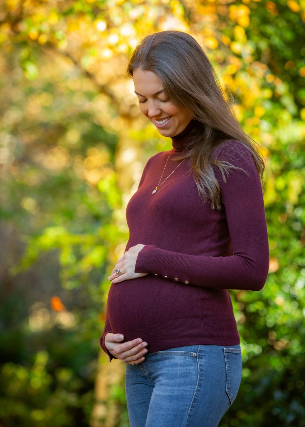 A smiling pregnant woman with long brown hair stands outdoors in sunlight, wearing a burgundy polo neck and blue jeans. She gently cradles her baby bump with both hands, surrounded by soft-focus greenery and autumn-coloured foliage in the background.