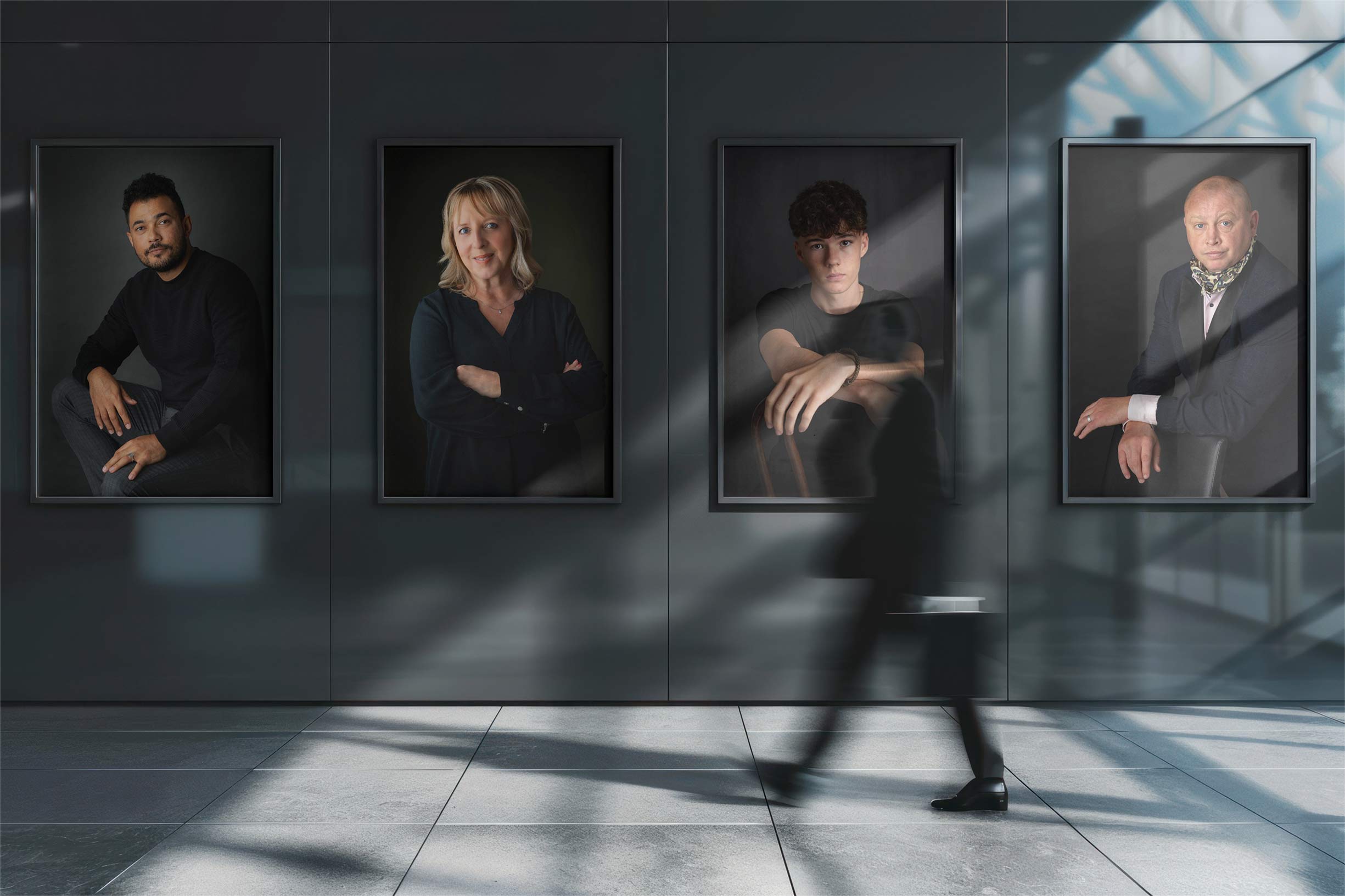 A blurred person walks past four large, framed portraits of three men and one woman hanging on a dark wall in a modern building with glass walls and sunlight streaming in, casting diagonal shadows on the floor and wall.