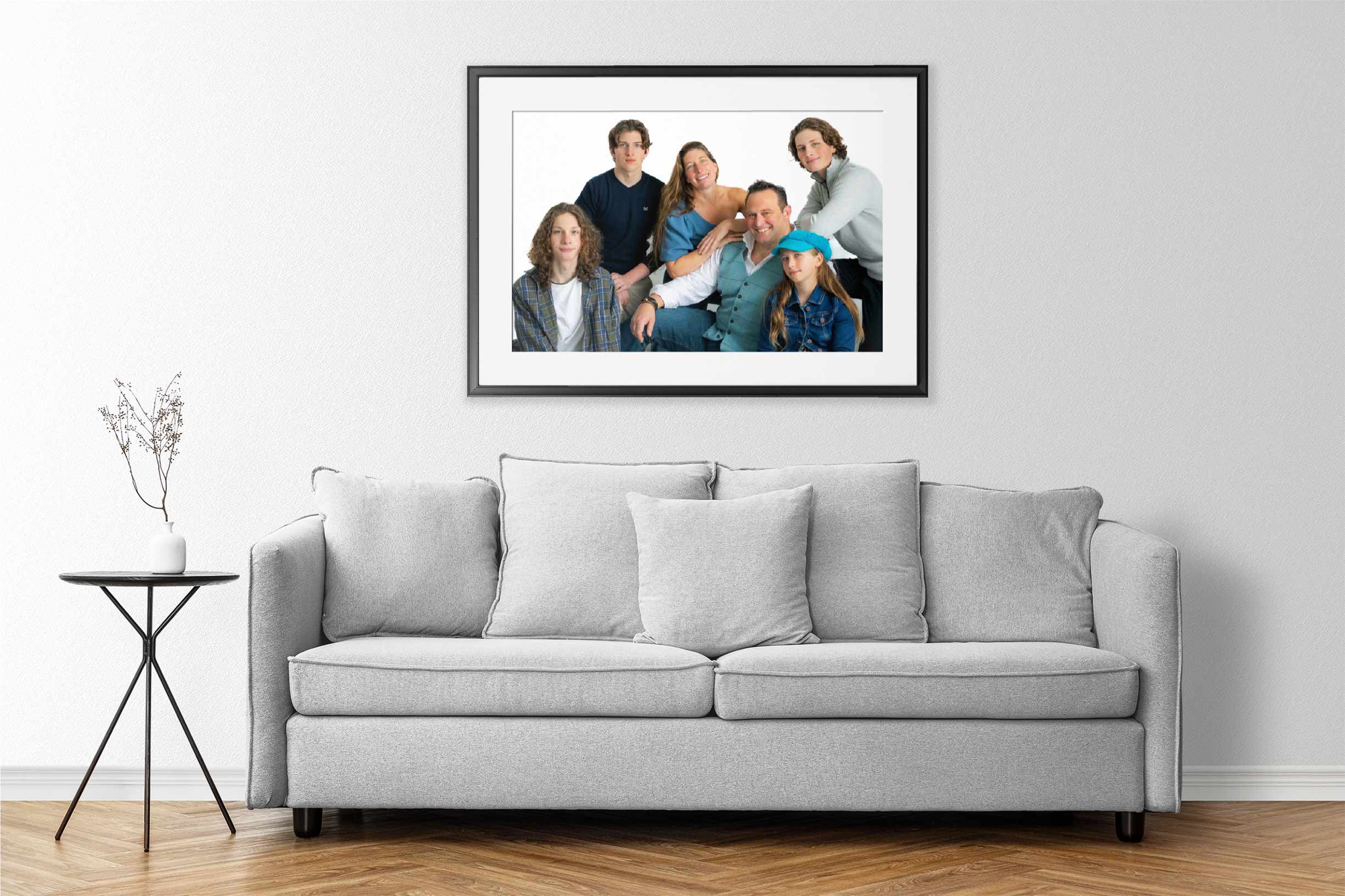 A modern living room with a light grey sofa, three cushions, a round black side table, and a family portrait on the wall above the sofa. The portrait shows six smiling people, including adults and children, posing together against a white background.