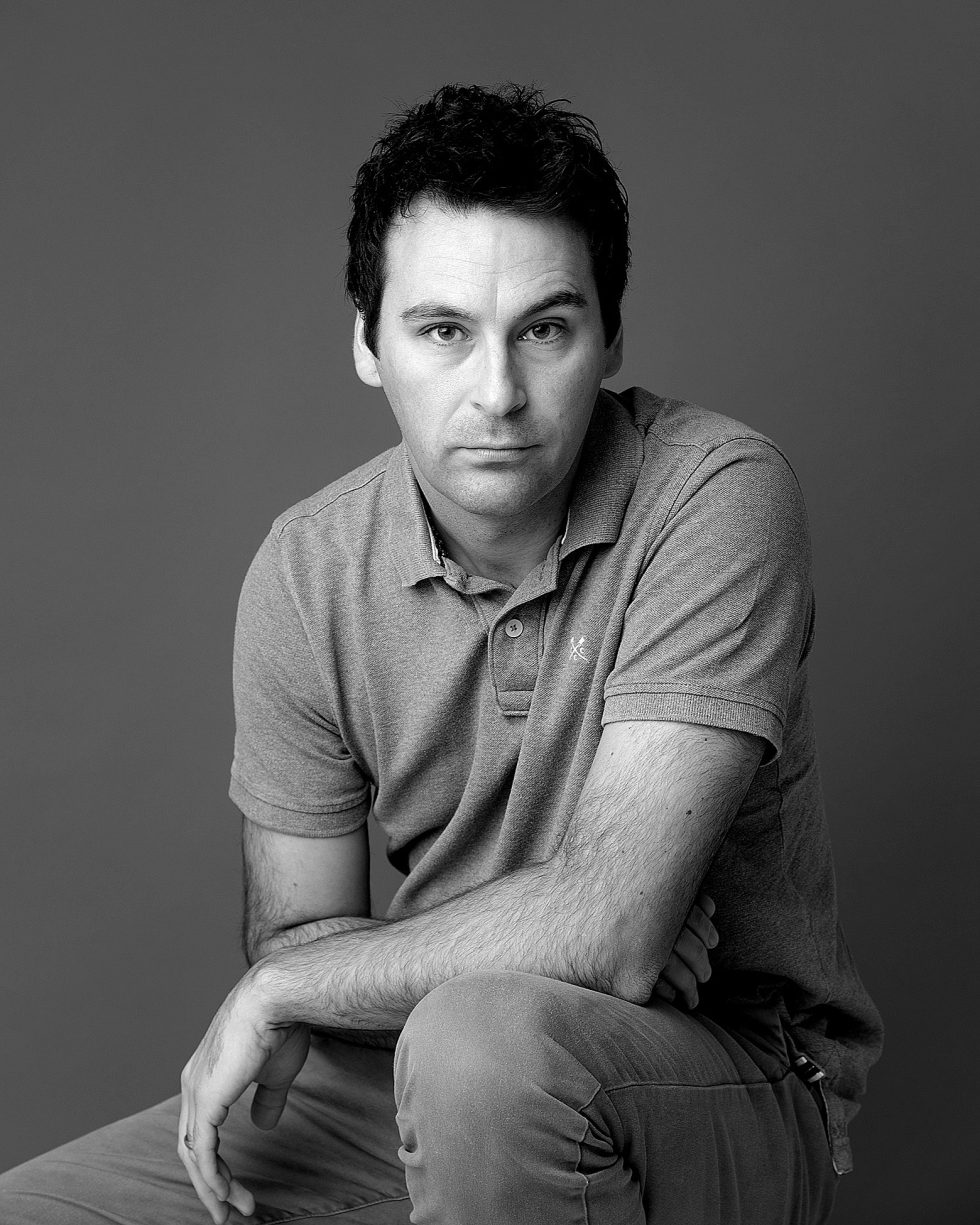 A man with short dark hair, wearing a short-sleeved polo shirt and light trousers, sits leaning slightly forward with one arm resting on his knee. He looks directly at the camera with a neutral expression. The background is plain and grey. The photo is in black and white.