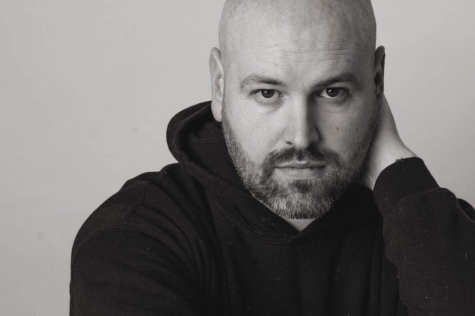 Black-and-white portrait of a bearded bald man in a dark hoodie. He looks directly at the camera with a serious expression, resting one hand on the back of his neck. The background is plain and light-coloured, emphasising his facial features.