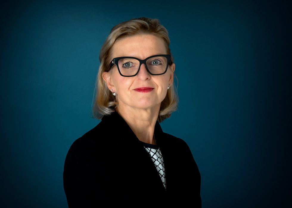 A woman with blonde hair, wearing black-framed glasses, stud earrings, and a black blazer over a white and black patterned top, poses in front of a dark blue background. She is looking confidently at the camera with a slight smile.