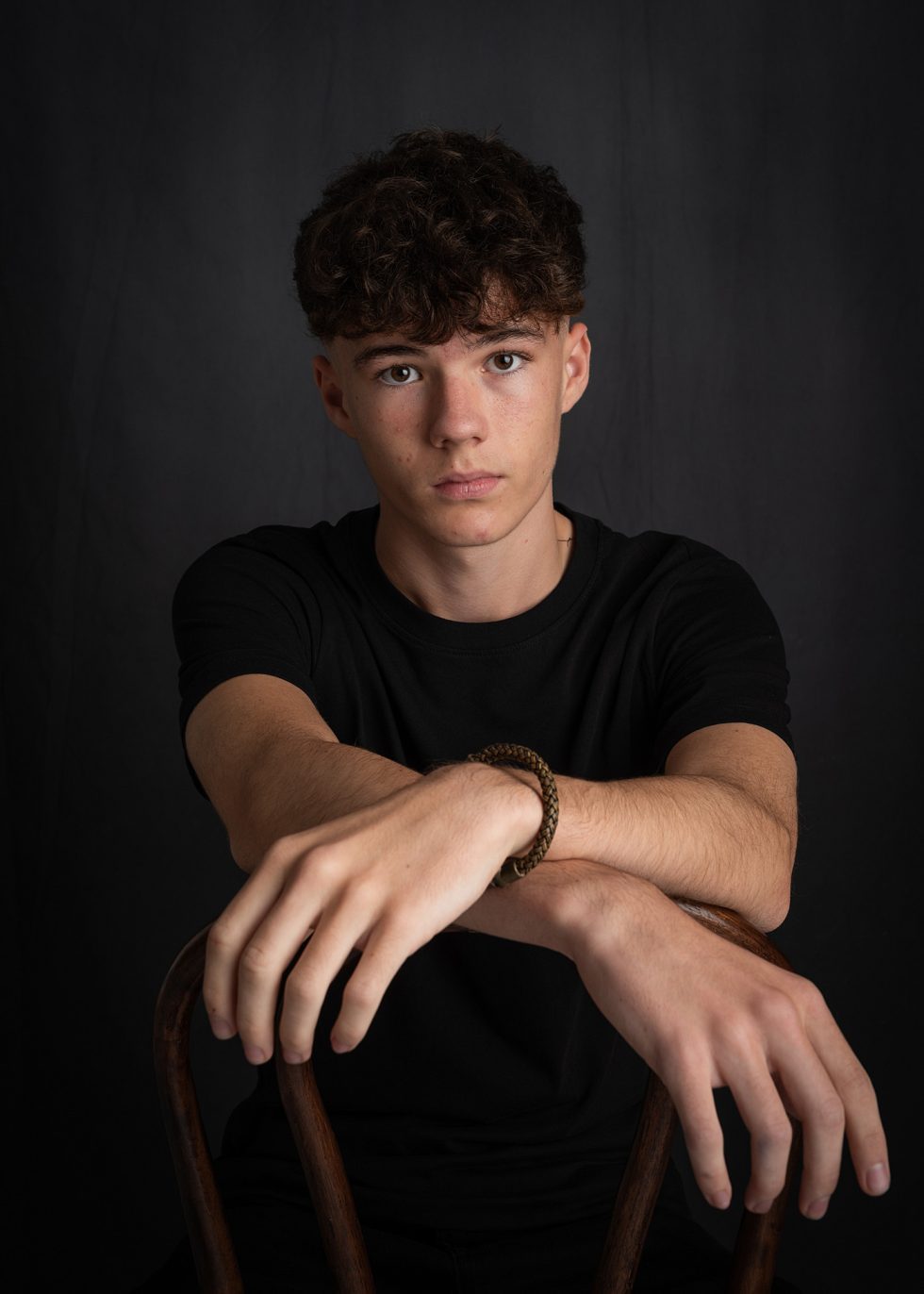 A young person with short curly brown hair and fair skin sits facing the camera against a dark background. They wear a black T-shirt and rest their arms on the back of a wooden chair, looking directly ahead with a neutral expression.