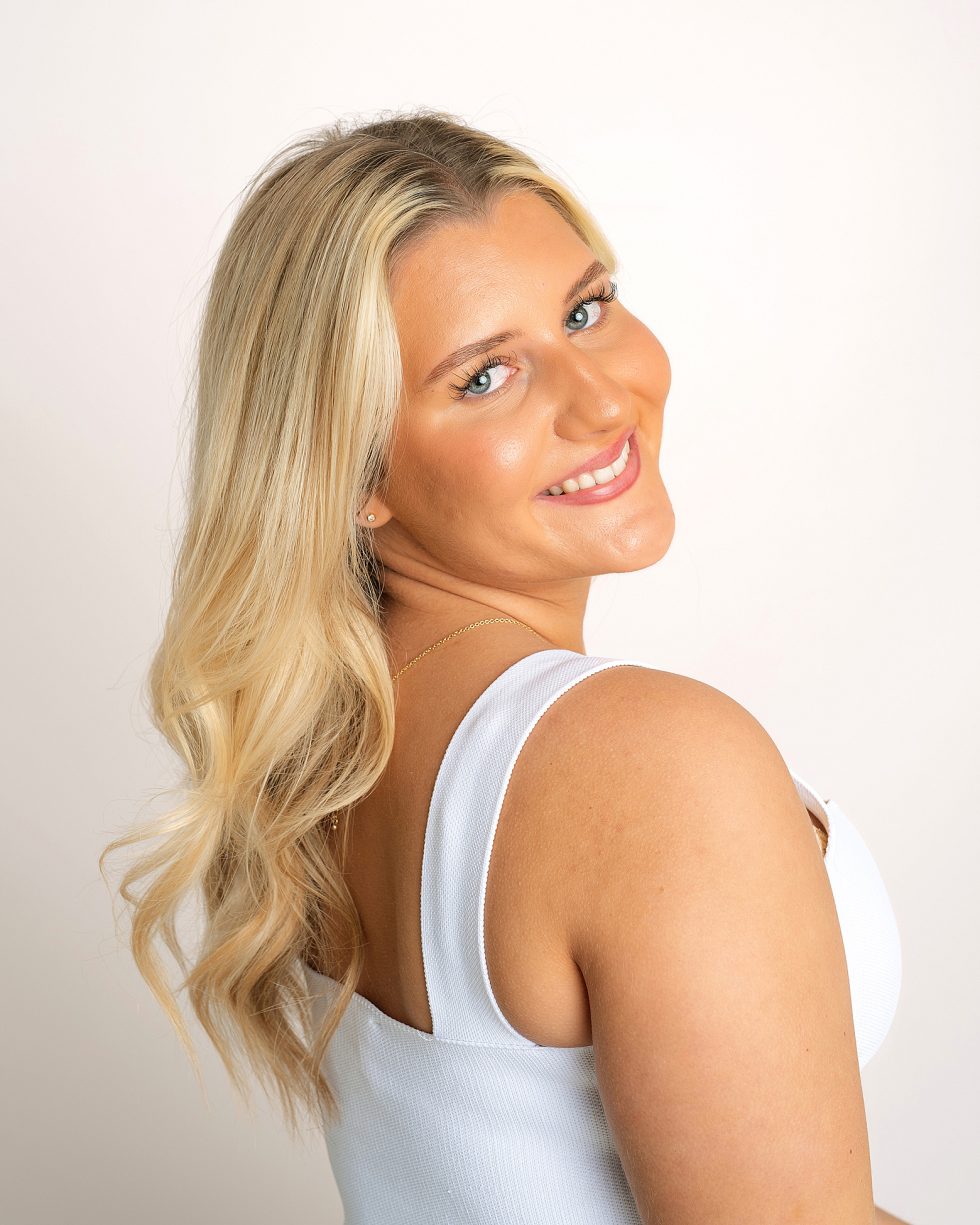 A woman with long, wavy blonde hair and blue eyes smiles over her shoulder at the camera. She is wearing a sleeveless white top and a gold necklace against a plain, light background. Her skin is tanned and her make-up is natural.