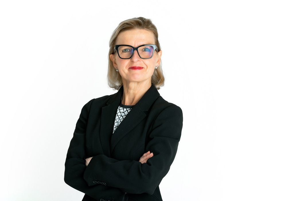 A confident woman with light skin, short blonde hair, and glasses stands against a white background. She wears a black blazer, a patterned shirt, and red lipstick, with her arms folded and a slight, professional smile.