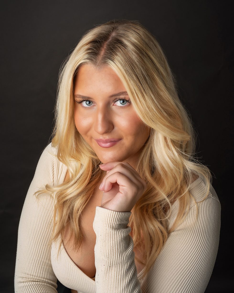 A young woman with long, wavy blonde hair poses against a black background. She wears a light beige, ribbed top with a deep neckline and smiles softly, resting her chin on her hand. Her make-up is natural, highlighting her blue eyes and warm skin tone.