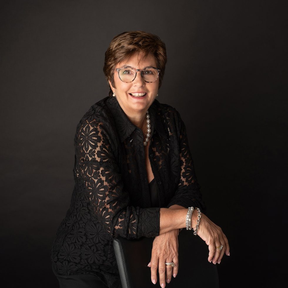 A smiling woman with short brown hair and glasses poses against a black background. She wears a floral-patterned black lace top, a pearl necklace, bracelets, and rings, and leans on the back of a black chair with her arms crossed.