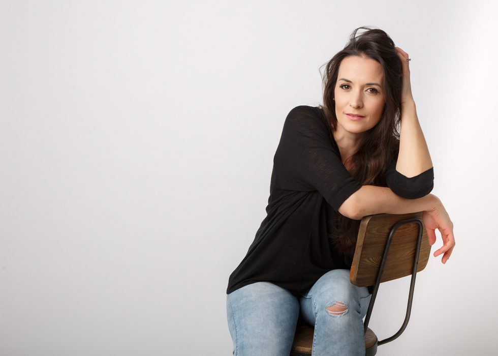 A woman with long dark hair, wearing a black top and ripped light blue jeans, sits sideways on a wooden chair. She rests one arm on the chair’s back and her head on her hand, looking calmly at the camera. The background is plain and light-coloured.