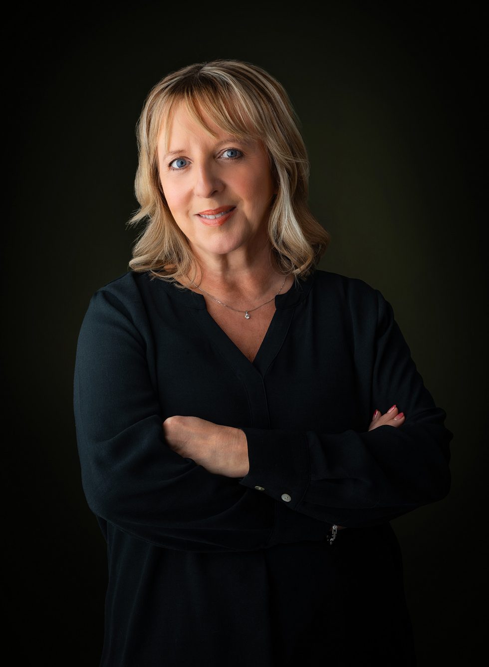 A woman with shoulder-length blonde hair and light skin stands against a dark background. She is wearing a long-sleeved black top and a delicate necklace, with her arms folded and a gentle, confident smile on her face.