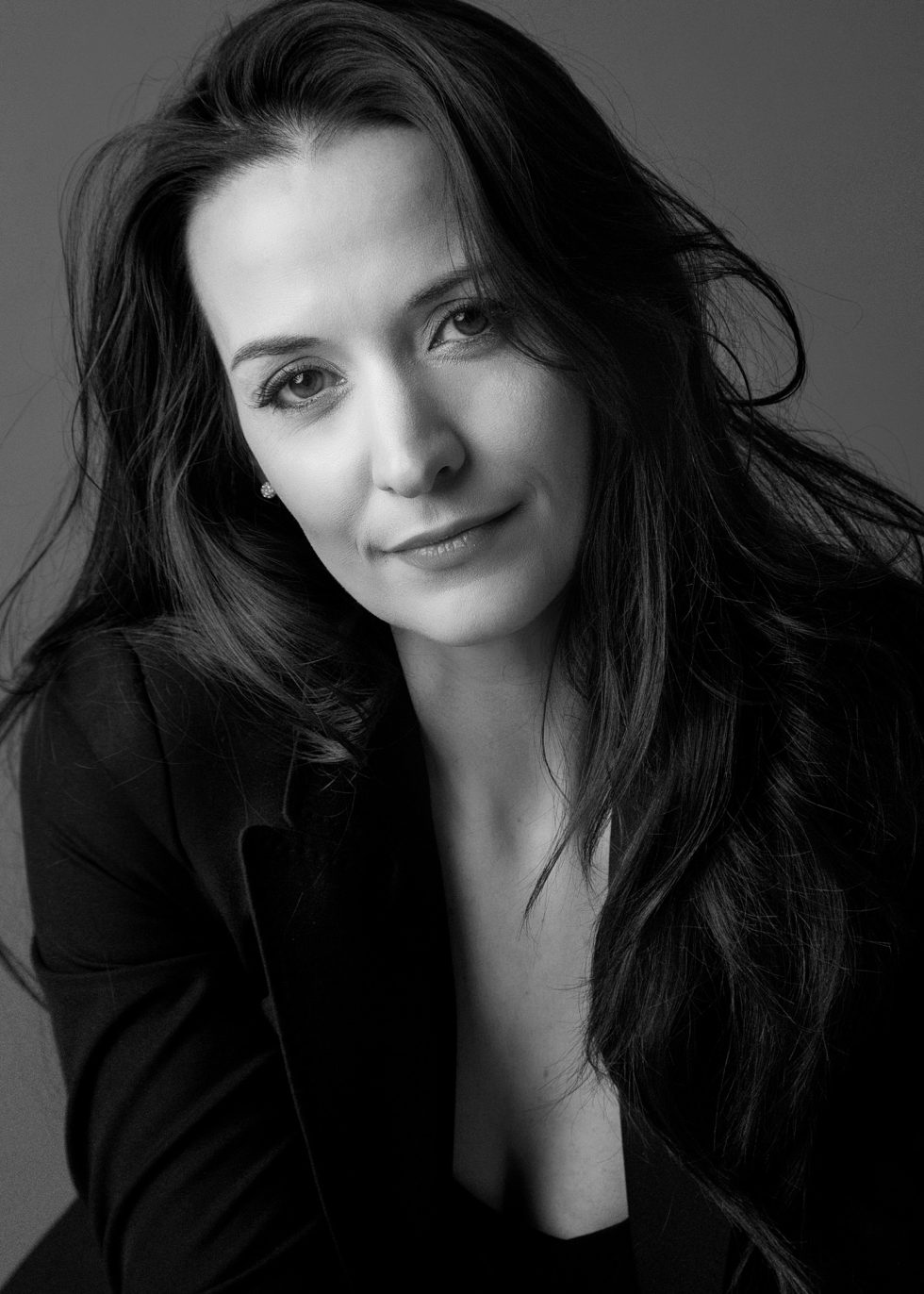 Black and white portrait of a woman with long dark hair, wearing a blazer over a top. She is facing the camera with a slight, natural smile and a relaxed expression. The background is plain and softly lit.