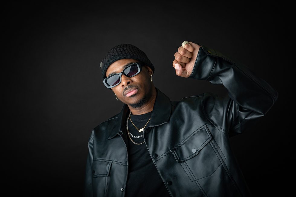 A man in a black leather jacket, black shirt, beanie, and sunglasses raises his right fist. He wears gold chains and hoop earrings, and stands against a dark background in a confident pose.