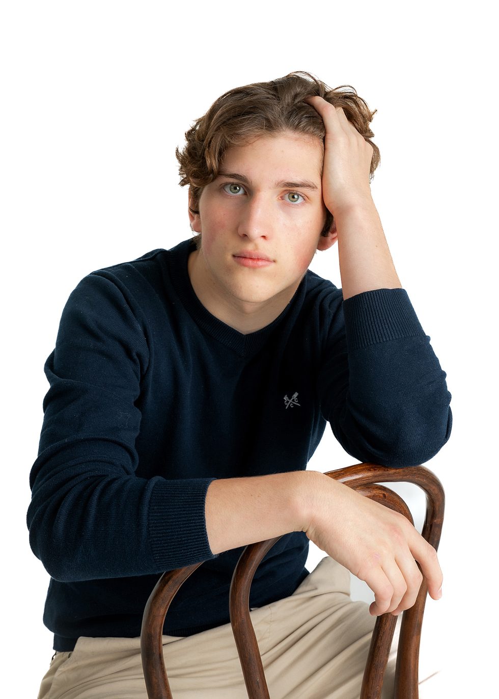 A teenage boy with wavy brown hair and blue eyes sits on a wooden chair, resting one hand on his head and the other on the chair’s back. He wears a navy jumper and beige trousers, looking directly at the camera with a neutral expression. White background.
