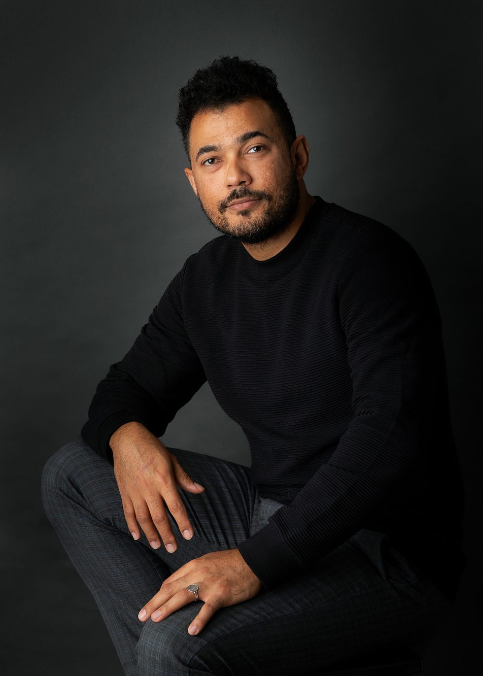 A man with curly dark hair and beard sits on a stool against a dark background. He wears a black jumper and grey checked trousers, resting one hand on his knee and the other on his leg, looking at the camera with a calm, slight smile.