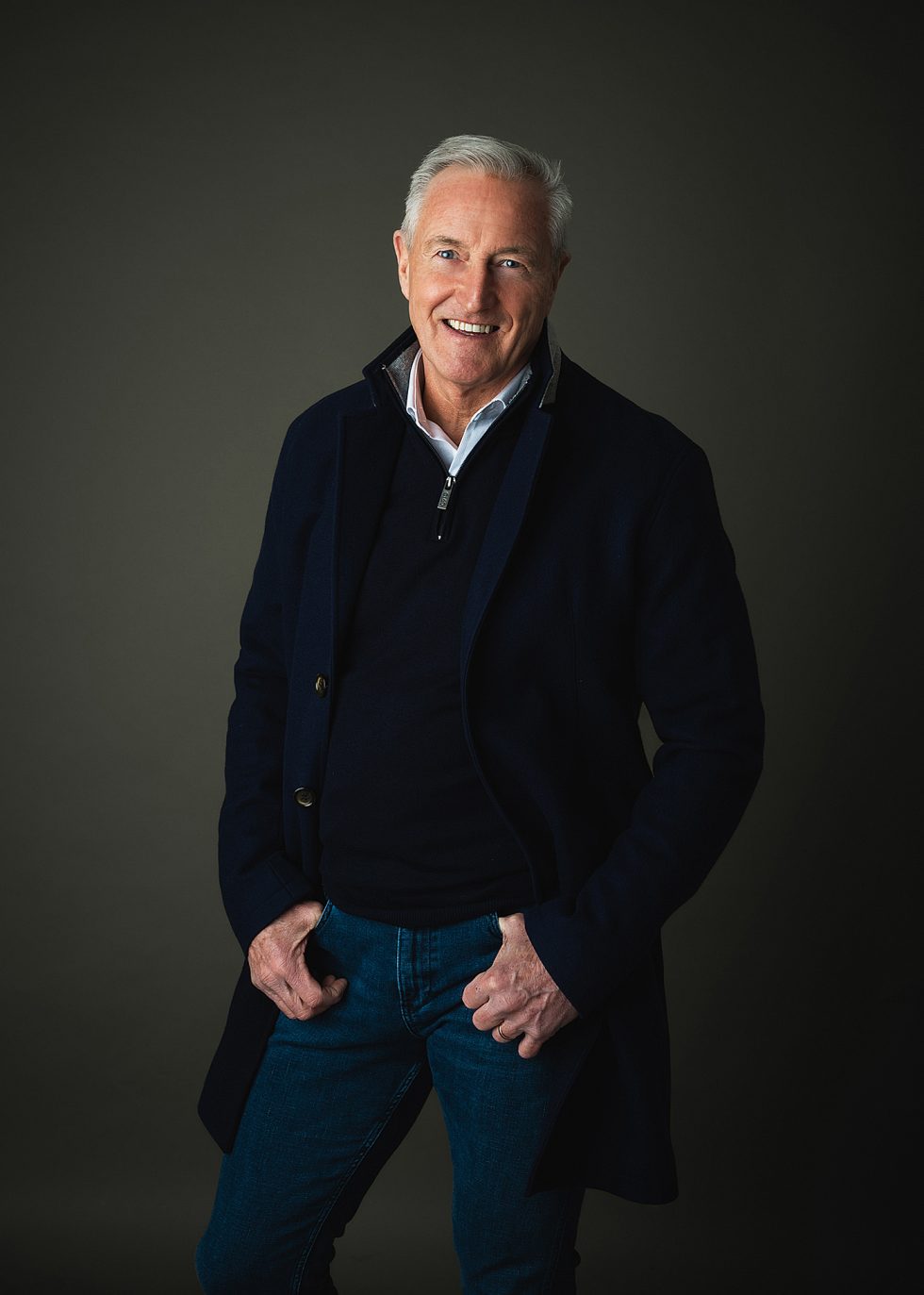 A smiling older man with short grey hair stands against a dark background. He wears a navy coat over a dark jumper and white collared shirt, along with blue jeans. His hands rest casually in his pockets and he looks directly at the camera.