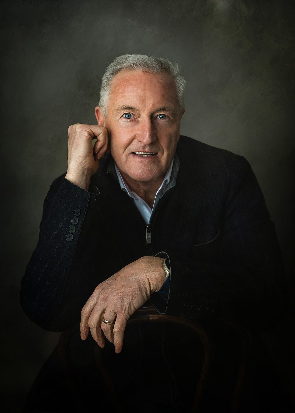 An older man with short grey hair and blue eyes poses, smiling gently. He wears a dark blazer over a light shirt and a watch on his left wrist. He rests one arm on a chair’s back and his chin on his other hand, with a muted green-grey background.
