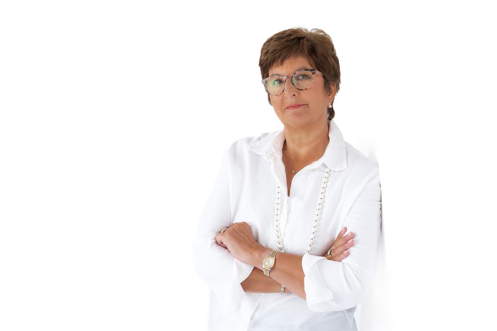 A middle-aged woman with short brown hair and glasses stands against a white background. She wears a white button-up shirt, a long beaded necklace, and a watch, with her arms crossed and a slight smile on her face.