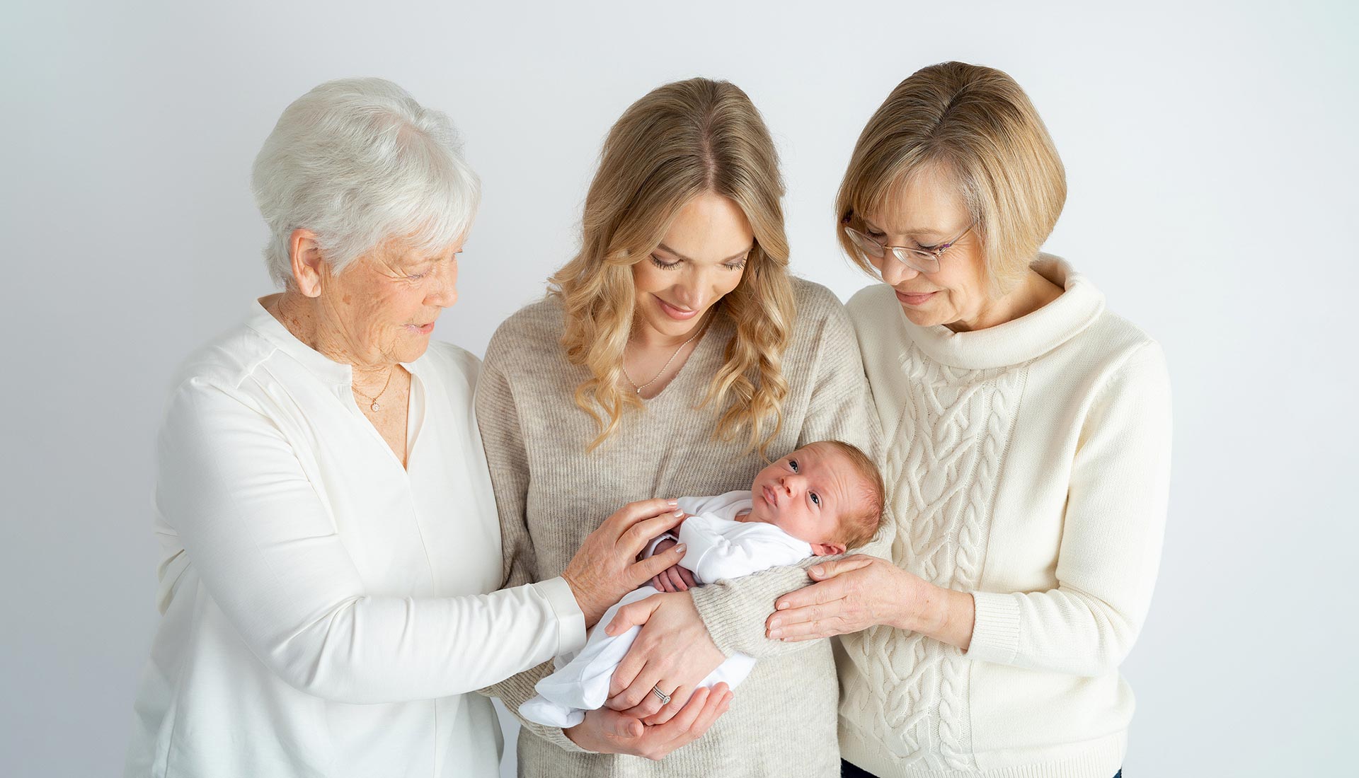 Three women of different generations, all with fair hair and wearing light-coloured jumpers, gather closely and smile while looking at a newborn baby in the centre. The baby is swaddled in white and cradled gently in the middle woman's arms.