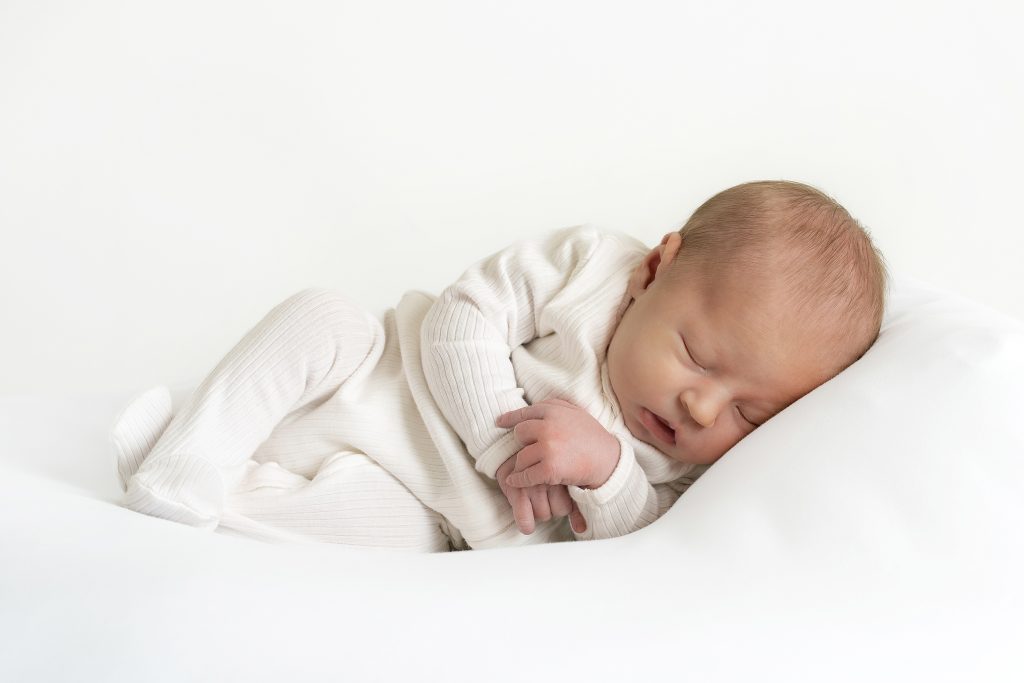 A newborn baby sleeps peacefully on a white pillow, dressed in a light cream-coloured, long-sleeved babygrow. The baby is lying on its side with eyes closed, hands gently clasped, and a calm, serene expression, against a plain white background.