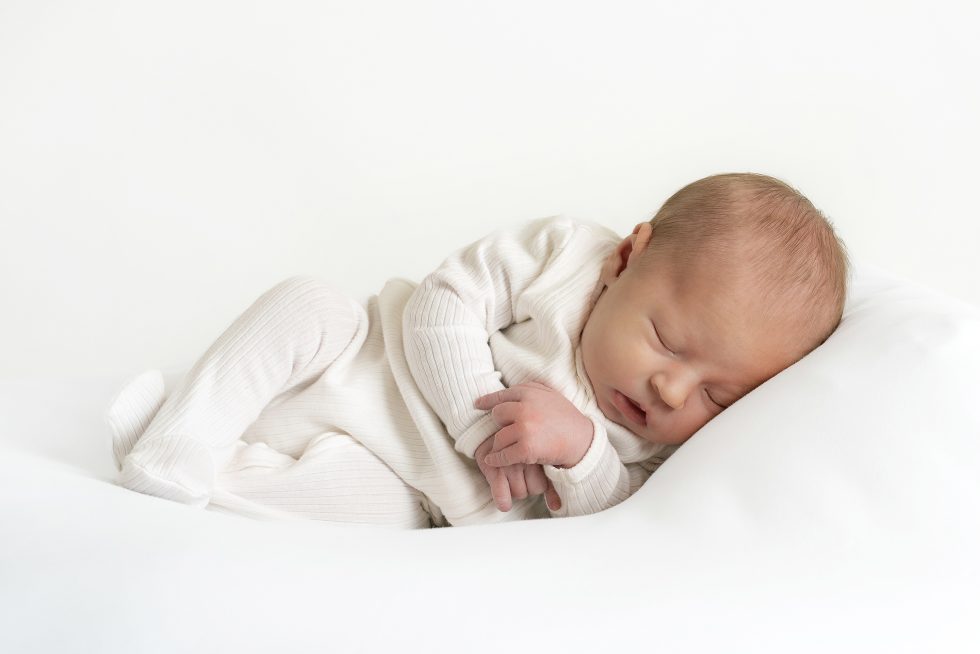 A newborn baby sleeps peacefully on a white pillow, dressed in a light cream-coloured, long-sleeved babygrow. The baby is lying on its side with eyes closed, hands gently clasped, and a calm, serene expression, against a plain white background.