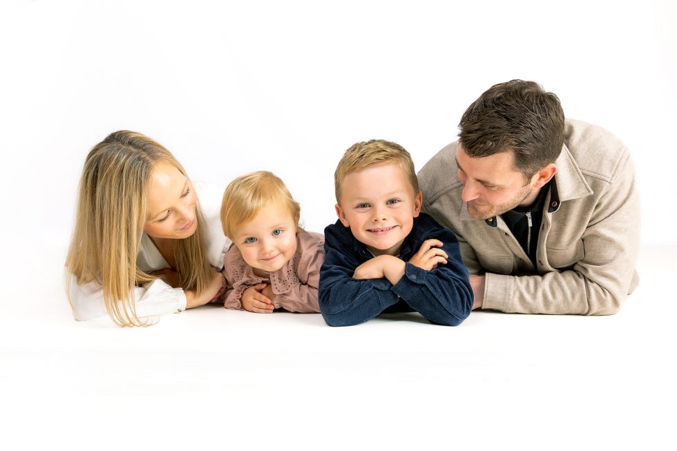 A smiling family of four lies on the floor facing the camera against a white background. Two adults, a woman and a man, are on the outer sides; between them are two young children, one toddler and one slightly older child, both smiling warmly.