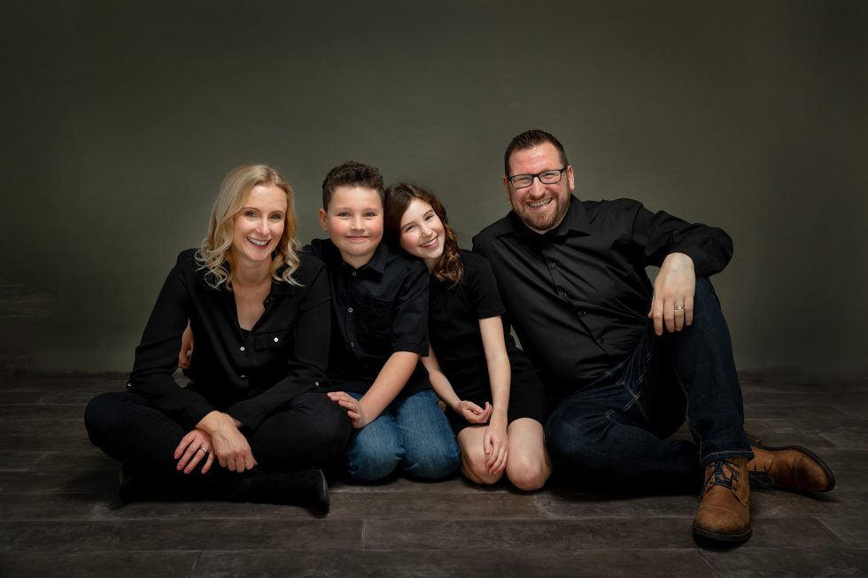 A smiling family of four sits close together on a dark wooden floor against a grey backdrop. The parents wear black tops; the mother sits cross-legged and the father has his arm round their son and daughter, who are sitting in the middle, all looking happy.