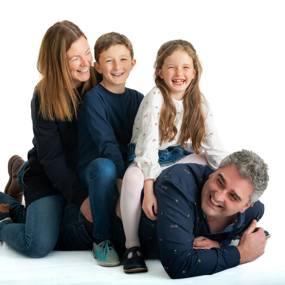A joyful family of four poses playfully, stacked on top of each other. The mother and son are at the back, the daughter sits in front, and the father lies at the bottom, smiling. All are casually dressed and laughing against a plain white background.
