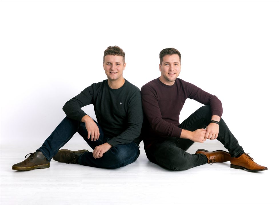 Two men sit on a white floor against a white background, both smiling at the camera. The man on the left wears a dark jumper, jeans, and brown shoes. The man on the right wears a maroon jumper, black jeans, and tan dress shoes.