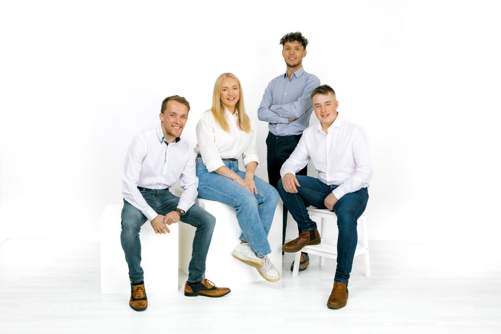 Four young adults pose in a bright studio with a white background. Two men sit on white cubes, a woman sits between them, and another man stands with arms crossed. They all wear casual attire: jeans, white shirts, and brown shoes. They are smiling at the camera.