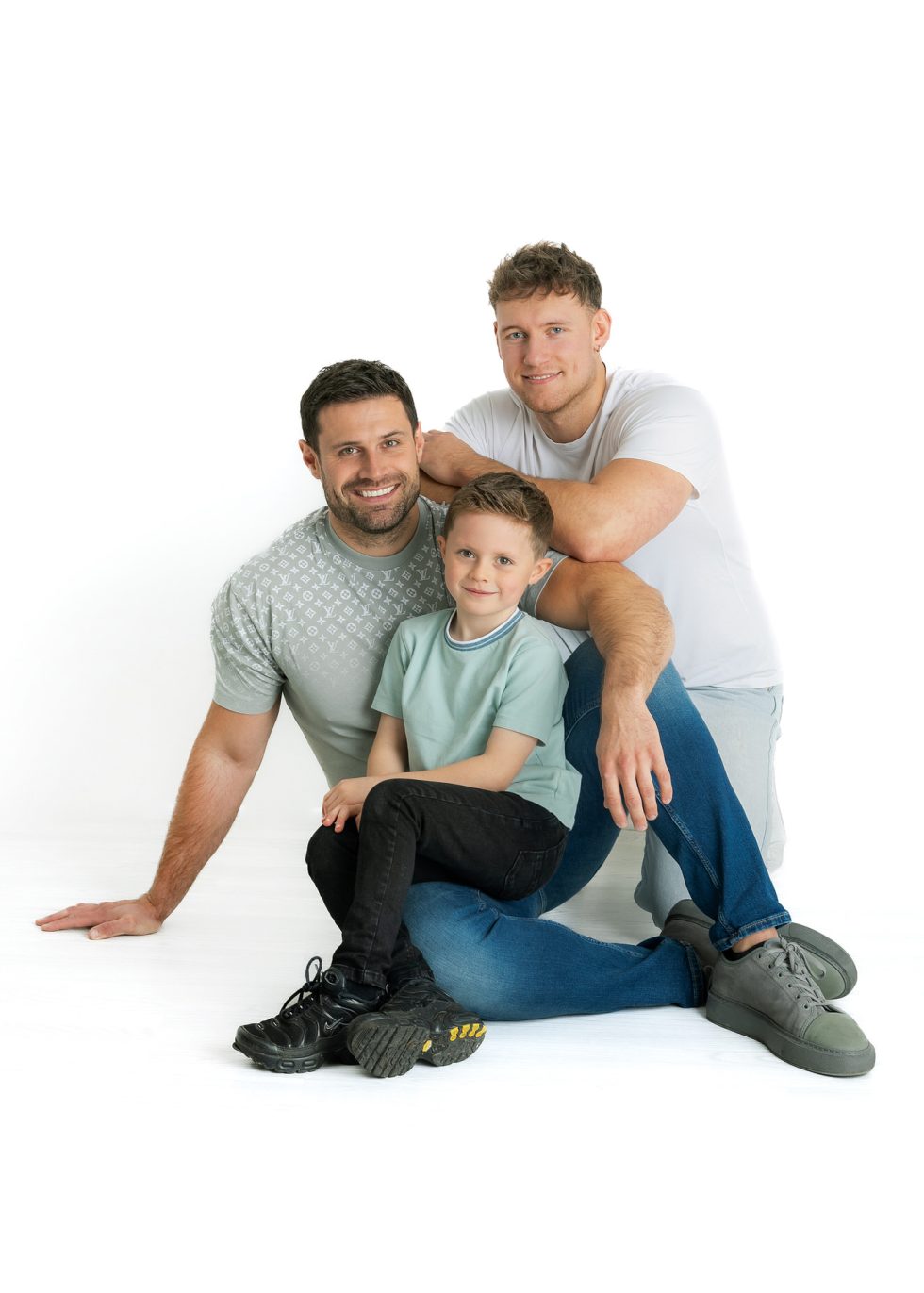 Three people pose in a studio against a white background: two men sit on the floor, smiling, with a young boy sitting in front of them. The men wear casual T-shirts and jeans; the boy has short hair, a light green shirt, black trousers, and trainers.