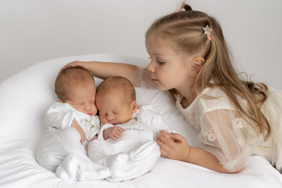 A young girl with long blonde hair and a white dress gently rests her hand on the heads of two newborn babies sleeping side by side on a white cushion. The babies are dressed in matching white outfits. The background is plain and light.